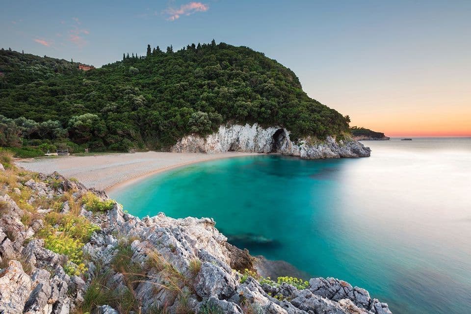 Una cala de arena con aguas tranquilas y turquesas, resguardada por un acantilado boscoso con una cueva marina al atardecer.