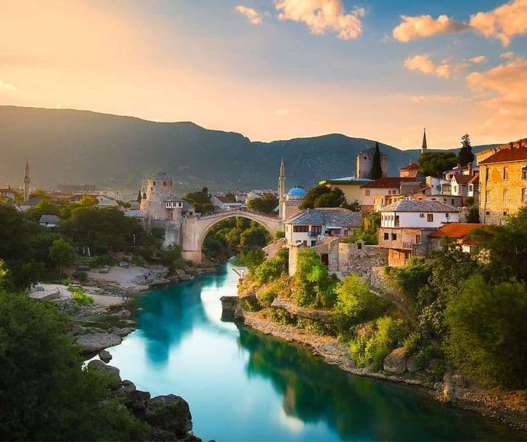 Un vecchio ponte ad arco in pietra attraversa un fiume turchese che scorre attraverso una città storica con montagne sullo sfondo al tramonto.