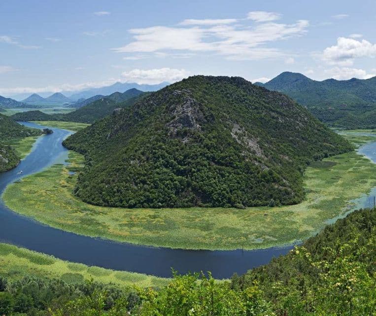 Un ampio fiume forma un'ansa a ferro di cavallo intorno a una grande collina boscosa, con rigogliose sponde verdi e montagne sullo sfondo.