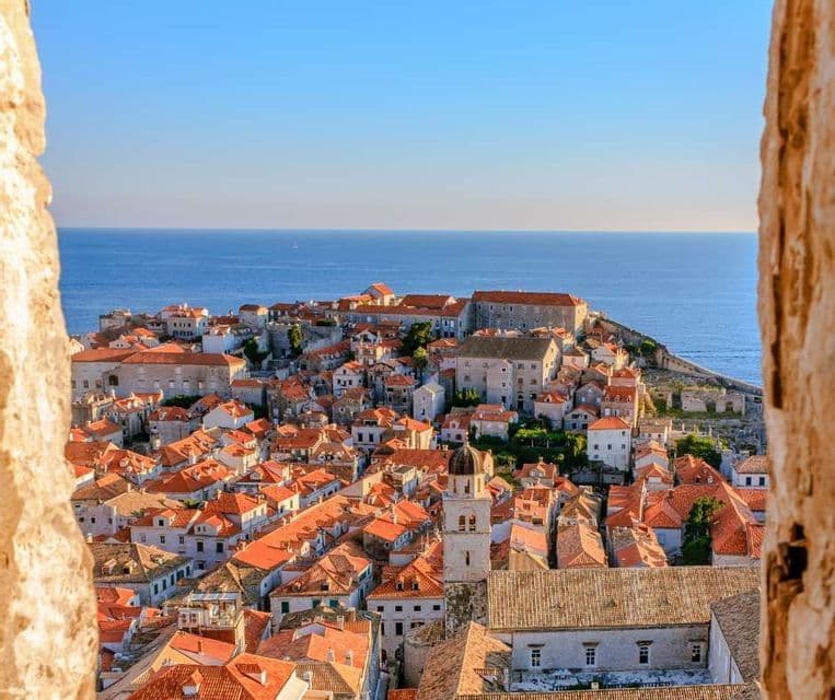 Una vista dall'alto di una città costiera con tetti in terracotta, scattata tra due mura di pietra e affacciata sul mare blu.