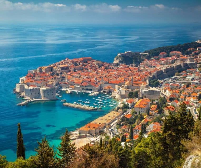 Una vista panoramica di una città costiera murata con tetti in terracotta, circondata da un mare blu calmo e un porto pieno di barche.