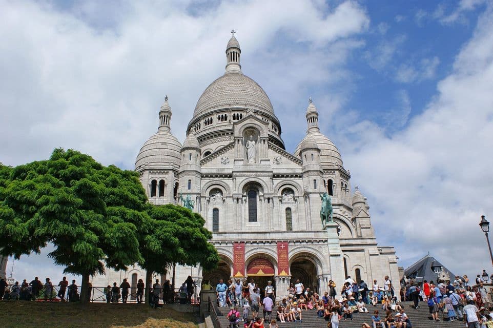 Le persone si radunano sui gradini di pietra fuori da una grande basilica dalla cupola bianca sotto un cielo blu parzialmente nuvoloso