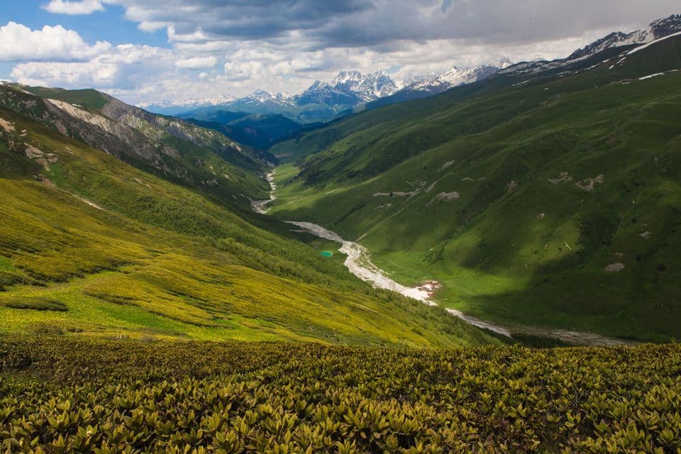 Un valle montana verde con un río serpenteante que se dirige hacia picos nevados bajo un cielo parcialmente nublado.