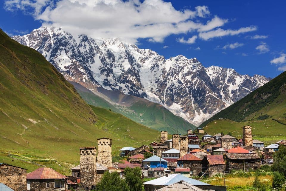 Antiguas torres de piedra y casas en un pueblo de montaña enclavado bajo enormes picos nevados bajo un cielo azul.