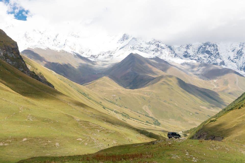Una furgoneta oscura aparcada en una colina cubierta de hierba con vistas a un amplio valle y picos de montañas nevadas al fondo.