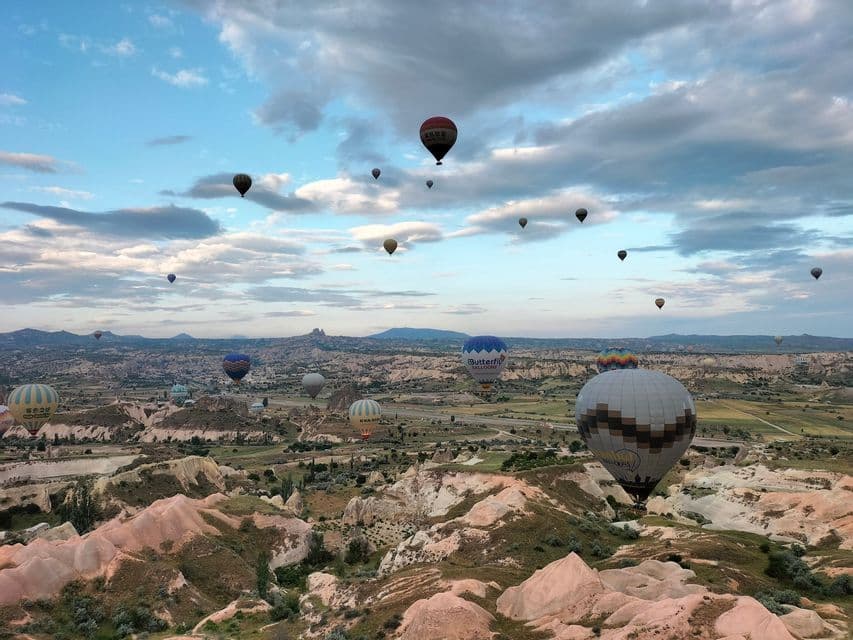 Numerosos globos aerostáticos flotando sobre un vasto paisaje de valle rocoso bajo un cielo parcialmente nublado.