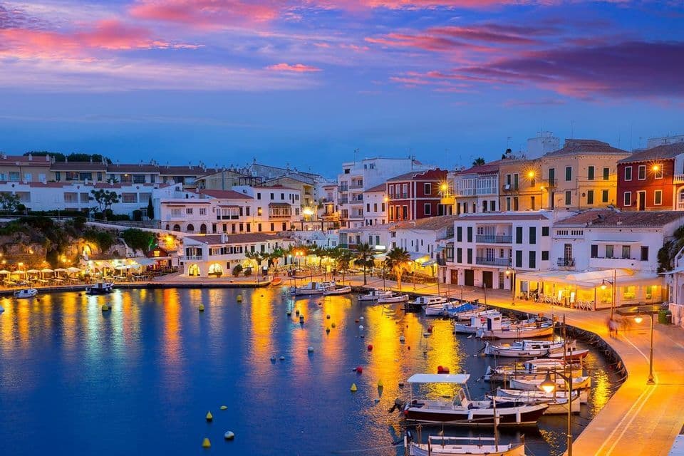 A harbor town at dusk, with lights from buildings and boats creating colorful reflections on the calm water.