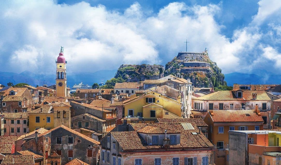 A cityscape of terracotta-roofed buildings with a prominent bell tower and a fortress on a hill under a cloudy blue sky.