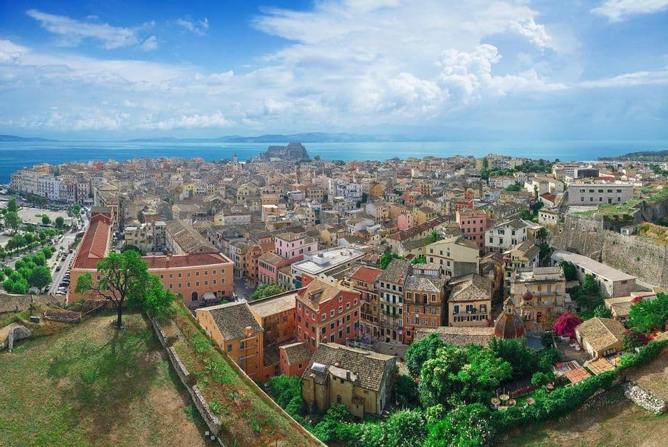 A high-angle view of a sprawling coastal city with colorful buildings and terracotta roofs, with the blue sea and a partly cloudy sky in the background.