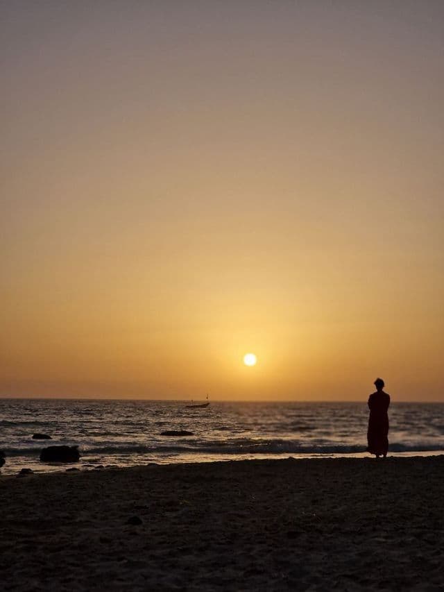 A person in silhouette stands on a sandy beach watching the sun set over the ocean, with a small boat in the distance.