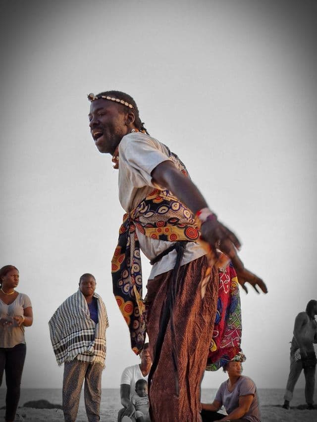 A man in colorful traditional clothing and a shell headband dances energetically outdoors for a group of onlookers.