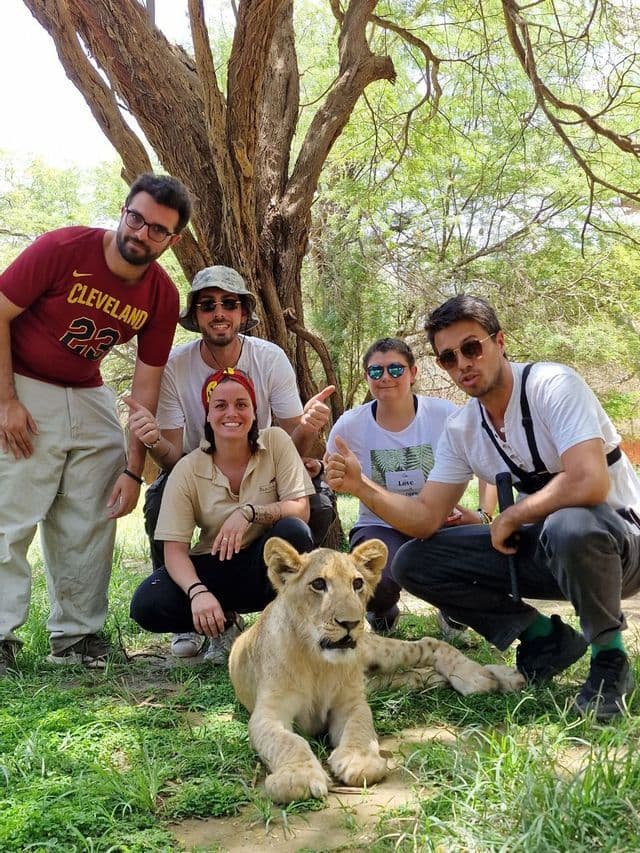 A WeRoad group trip of people posing for a photo with a lion cub lying on the grass under a large tree.