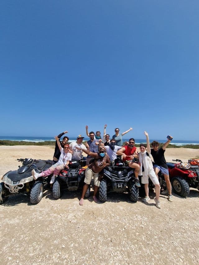 A WeRoad group trip posing for a photo on and around their quad bikes on a dirt path with the ocean in the background.