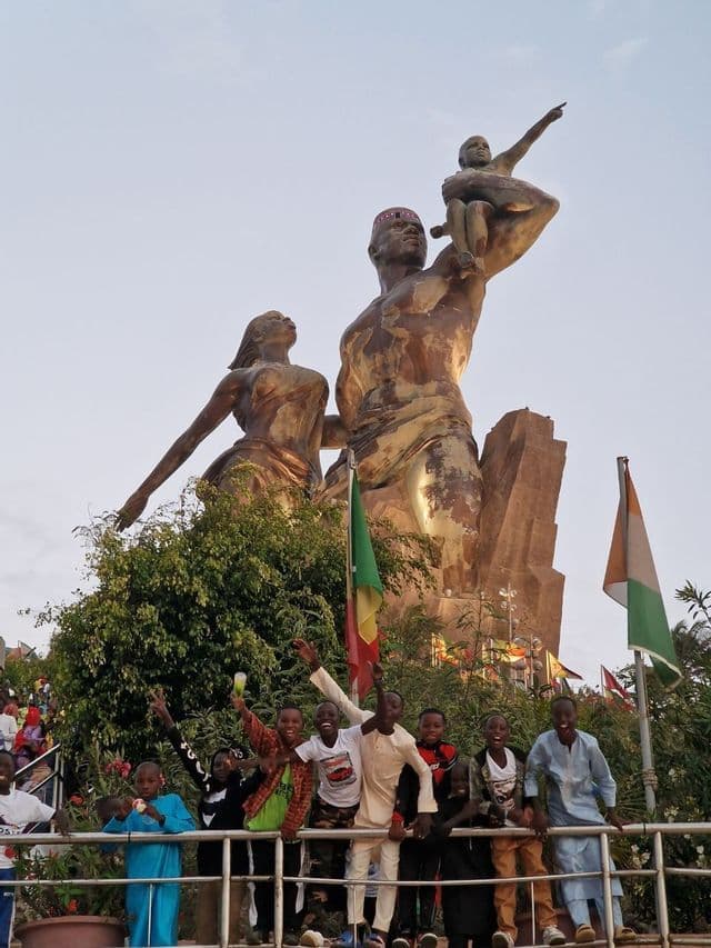 Un gruppo di ragazzi sorridenti posa per una foto davanti a una monumentale statua di bronzo di una famiglia contro un cielo pallido.