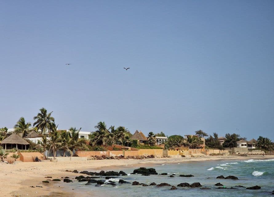 A coastal village with palm trees and traditional buildings sits on a sandy beach next to a rocky shoreline under a clear blue sky.
