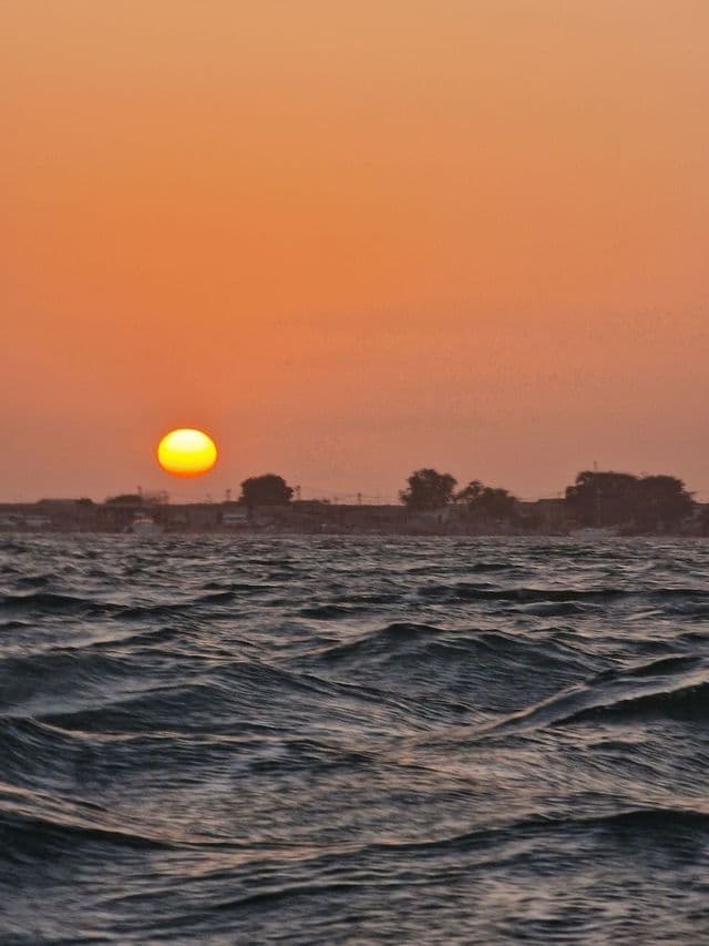 The sun sets over the choppy sea, casting an orange glow on the sky above a distant shoreline.