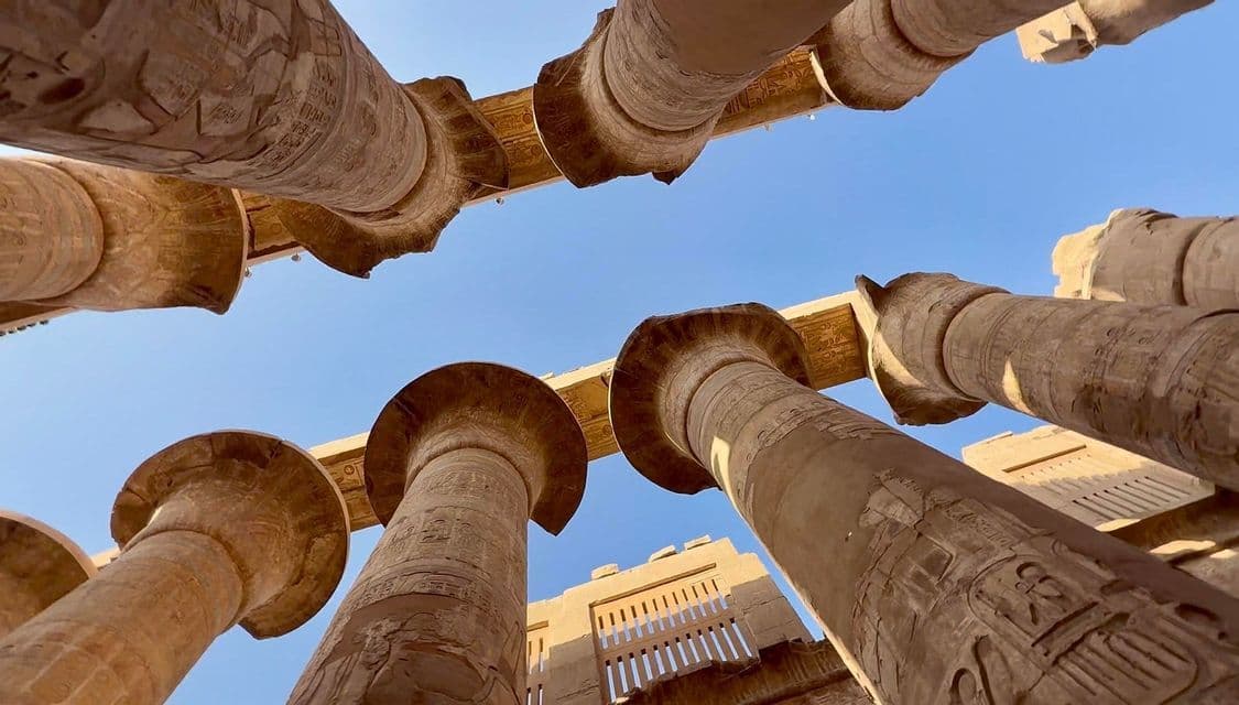 Inquadratura dal basso di antiche colonne di pietra scolpite con geroglifici che si ergono verso un cielo azzurro e limpido