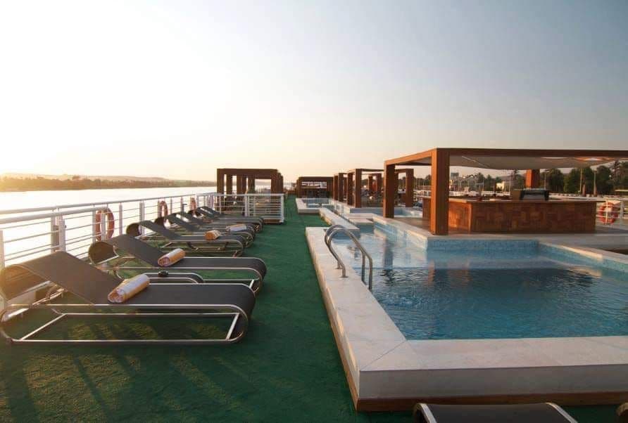 Lettini prendisole fiancheggiano una piscina su un ponte scoperto di una nave da crociera che si affaccia sull'acqua sotto un cielo limpido del mattino.