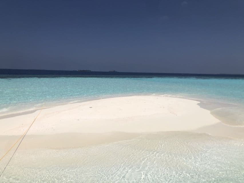 A white sandbar extends into clear turquoise water under a deep blue sky, with the dark ocean on the horizon.