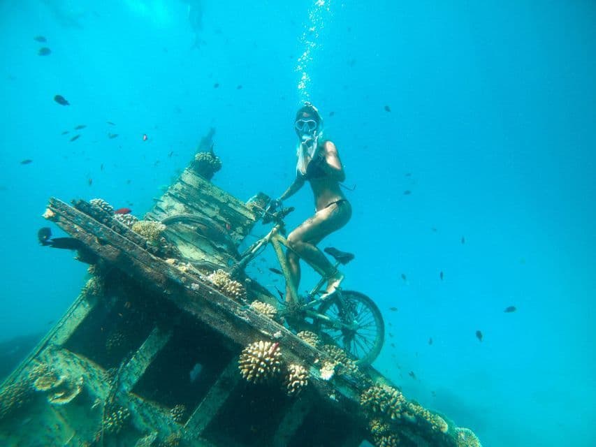 A woman in a bikini and diving mask sits on a bicycle on a sunken shipwreck, surrounded by blue water and small fish.