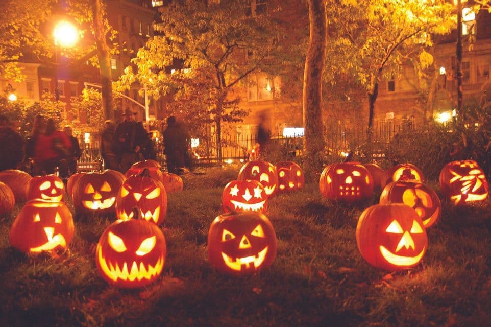 Un gruppo di jack-o'-lantern luminose con volti intagliati, sedute su un prato erboso in un parco di notte.
