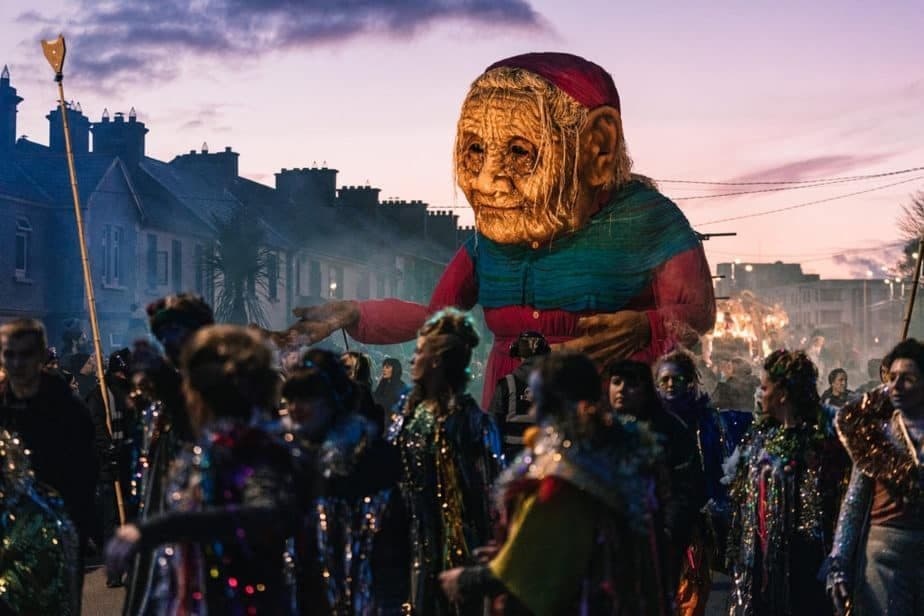 Un'enorme marionetta di un'anziana signora svetta su una folla in costumi festivi durante una sfilata di strada al tramonto.