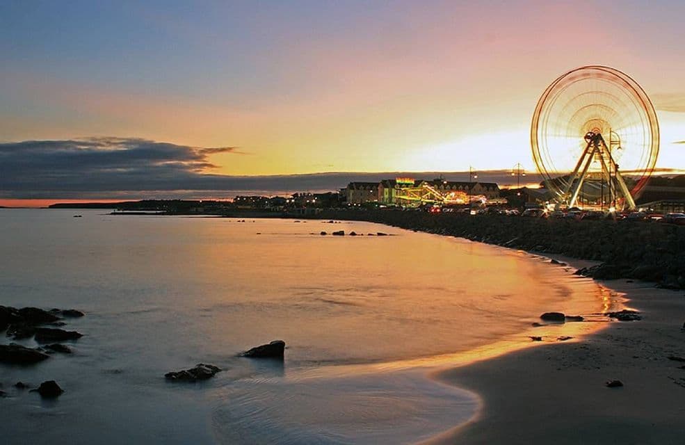 Una ruota panoramica illuminata si erge su una riva accanto a un mare calmo al tramonto, con il cielo colorato riflesso sull'acqua.