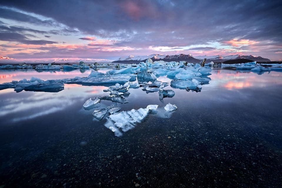 Icebergs float on calm water reflecting a sunset sky, with snow-capped mountains in the distance.