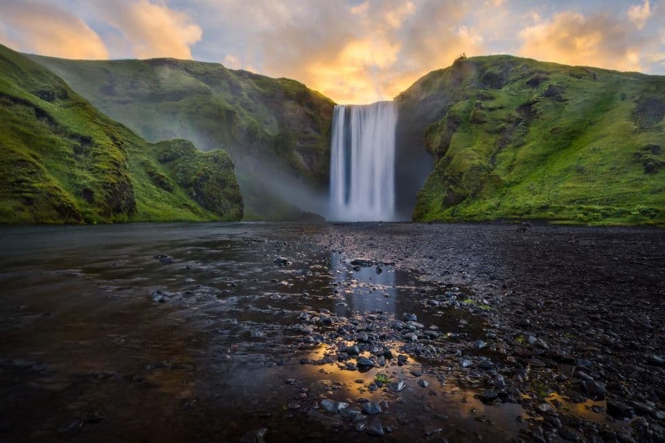 Un'ampia cascata scende tra due grandi scogliere verdi ricoperte di muschio in un letto di fiume roccioso al tramonto.