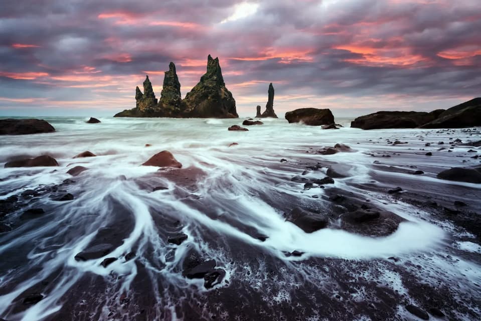 Long exposure of white waves washing over a black sand beach with jagged sea stacks in the ocean under a colorful, cloudy sky.