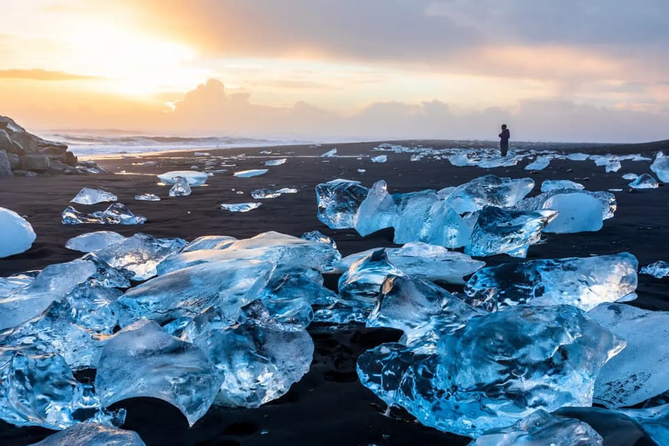 Grandes trozos de hielo cristalino están esparcidos en una playa de arena negra con una persona observando el océano al atardecer.