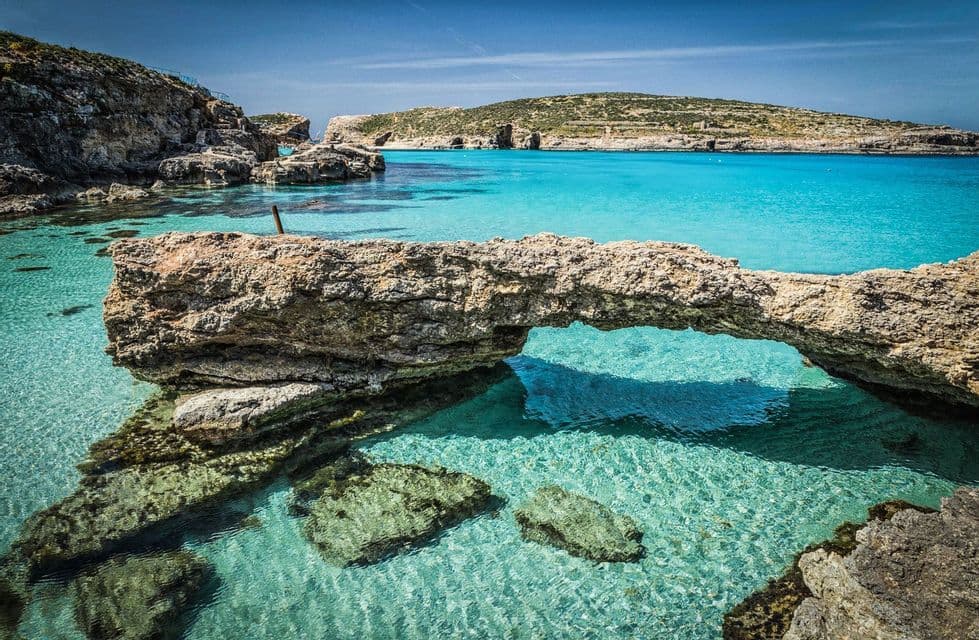 A natural rock arch over clear turquoise water in a rocky cove on a sunny day.
