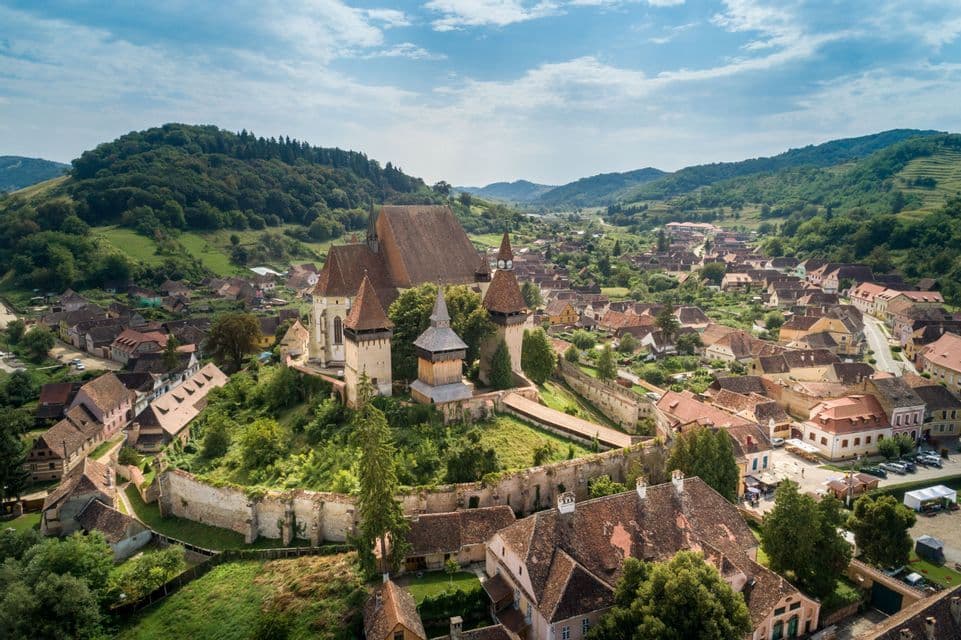 Una vista aerea di una storica chiesa fortificata su una collina, che domina un piccolo villaggio immerso in una valle verde.