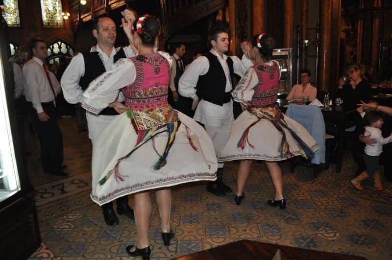 Deux couples vêtus de costumes traditionnels colorés interprètent une danse folklorique pour un public à l'intérieur d'un restaurant.