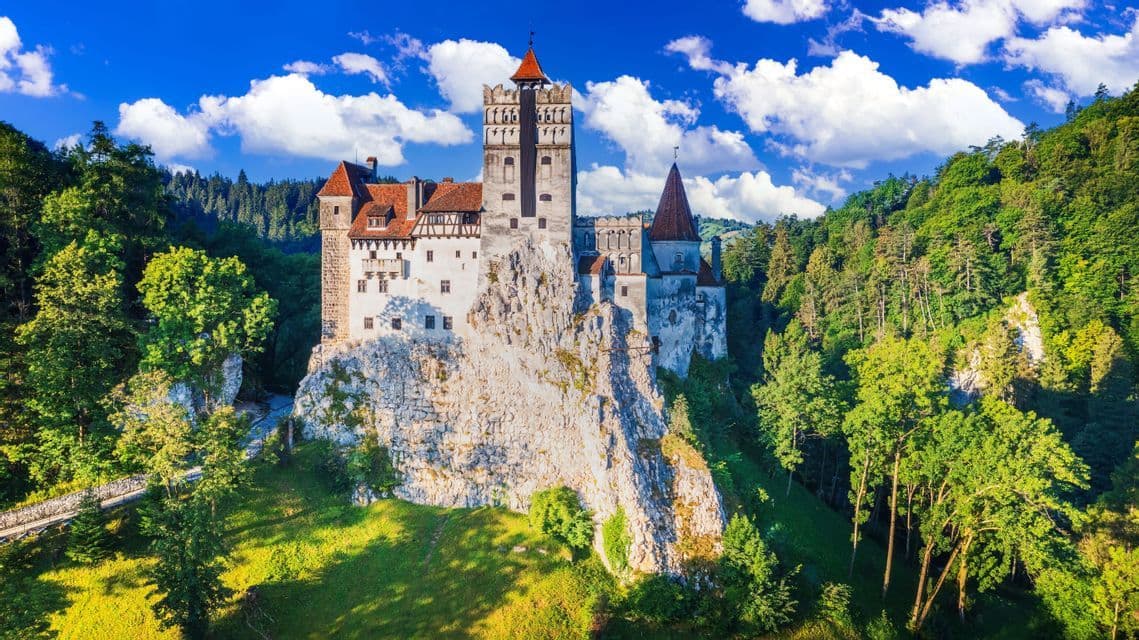 Un château de pierre aux toits rouges se dresse sur une falaise rocheuse, niché dans une forêt verte dense sous un ciel bleu partiellement nuageux.