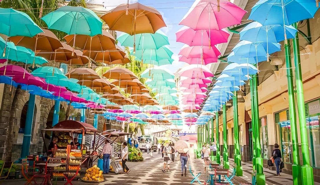 Decine di ombrelli colorati nelle tonalità del rosa, blu e marrone pendono su una strada pedonale assolata mentre le persone passeggiano.