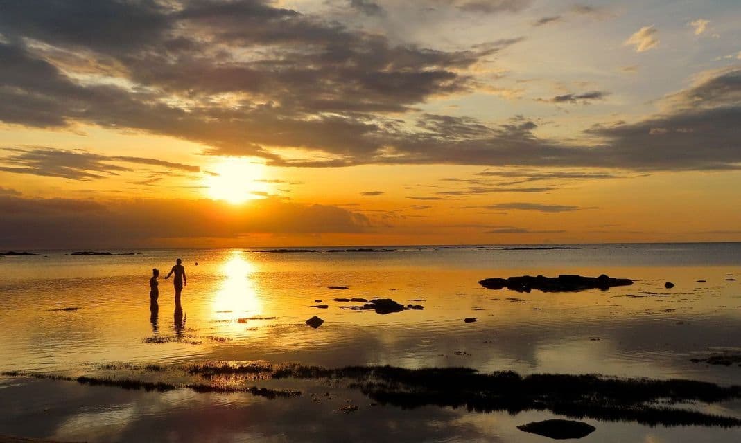 Sagome di due persone che si tengono per mano in un mare calmo durante un tramonto dorato, con il cielo riflesso sull'acqua.