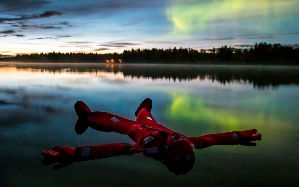 Una persona con un traje de supervivencia rojo flota de espaldas en un lago en calma, con la aurora boreal reflejada en el agua al atardecer.
