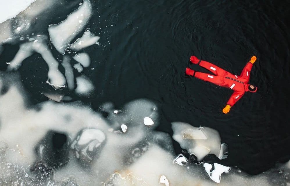 Vue aérienne d'une personne en combinaison de survie rouge flottant sur le dos dans une eau sombre et glacée.