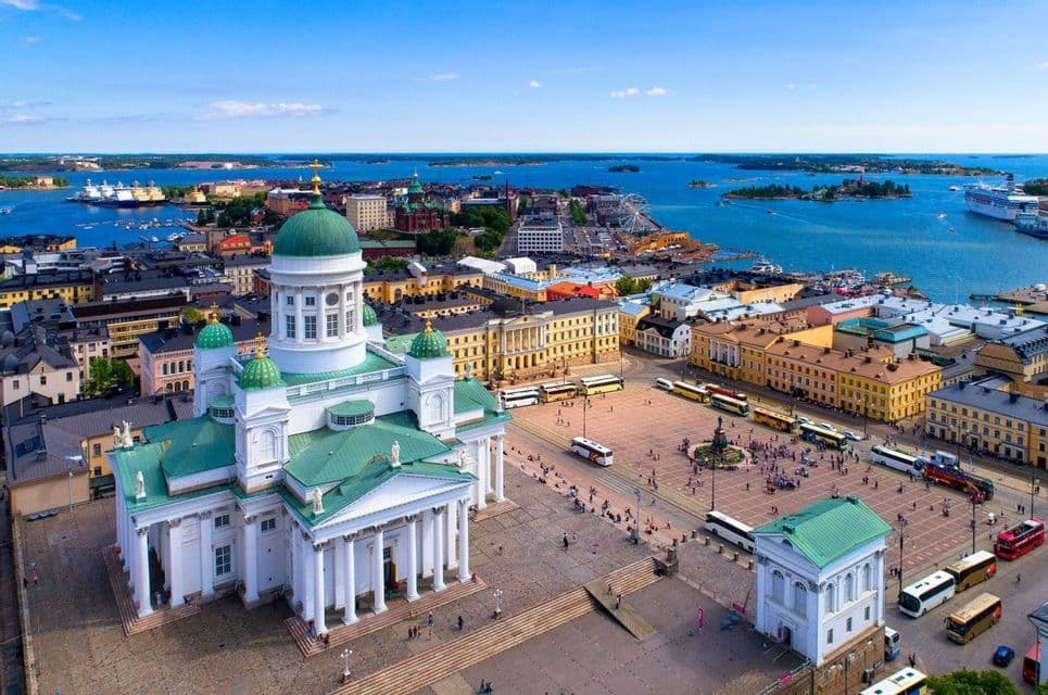 An aerial view of a large white cathedral with green domes overlooking a bustling city square and a bright blue sea with islands.