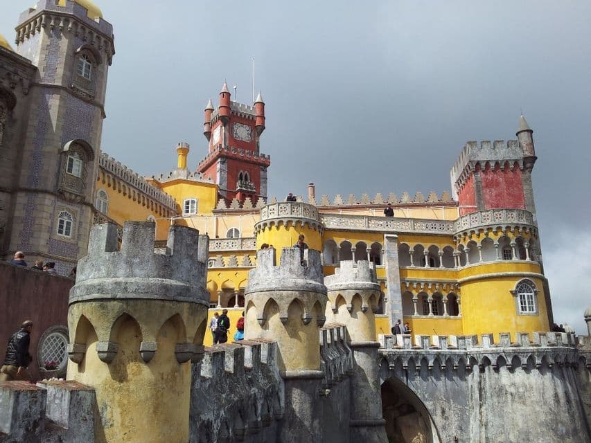 Un castillo colorido con torres amarillas y rojas, visto desde sus almenas de piedra donde la gente camina bajo un cielo nublado.