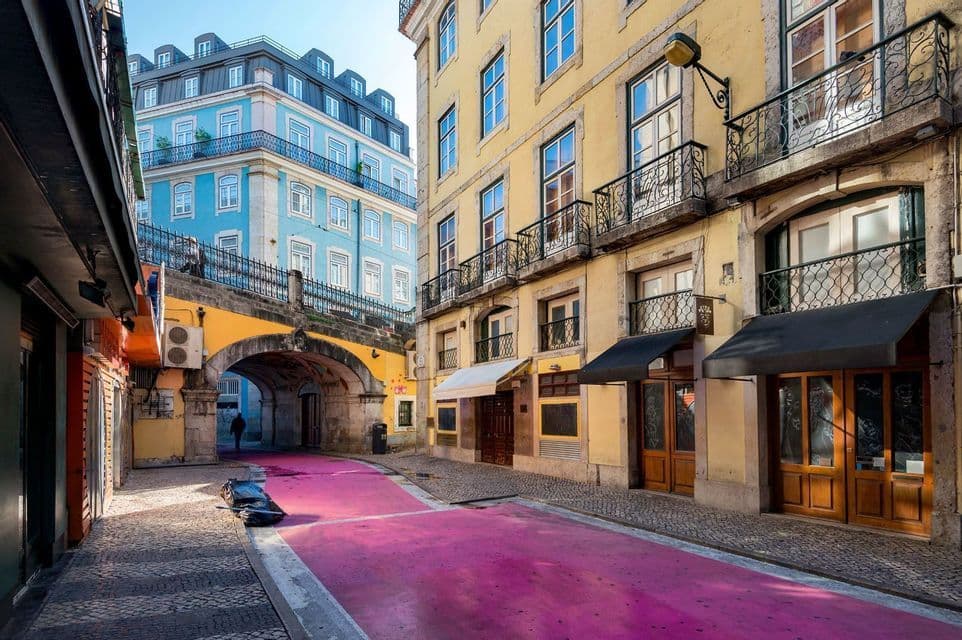 Eine schmale Stadtstraße mit einer leuchtend rosa Fahrbahn, die unter einem Steinbogen hindurchführt, gesäumt von farbenfrohen gelben und blauen Gebäuden.