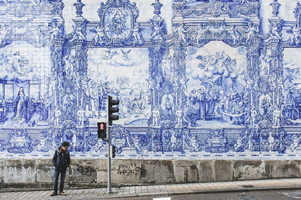 Eine Person steht auf einem Bürgersteig neben einer Ampel vor einer großen Wand, die mit kunstvollen blau-weißen Fliesen bedeckt ist.