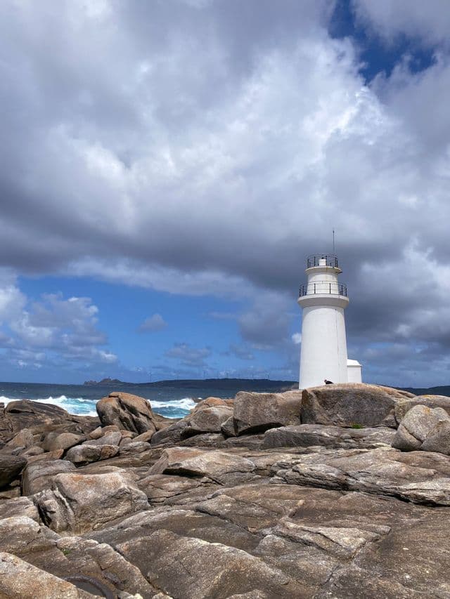 Un faro bianco si erge su una costa rocciosa accanto a un mare agitato sotto un cielo drammatico e nuvoloso.