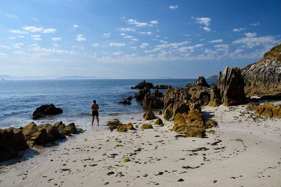 Una persona è in piedi sul bagnasciuga di una spiaggia sabbiosa e rocciosa, osservando l'oceano sotto un cielo azzurro con nuvole sparse.