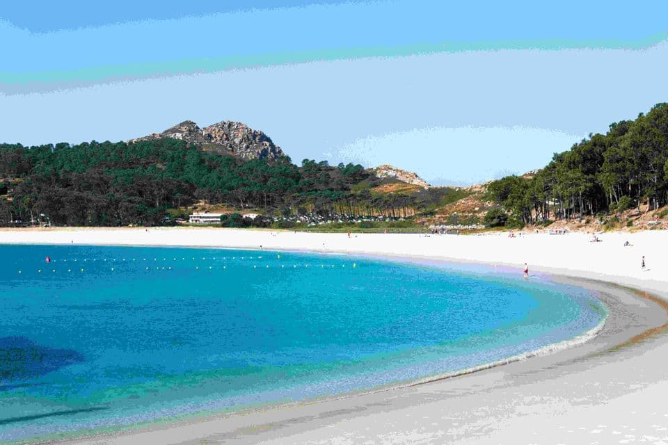 Una spiaggia di sabbia bianca e ampia si snoda lungo una baia turchese, con una collina boscosa che si erge sotto un cielo azzurro e limpido.