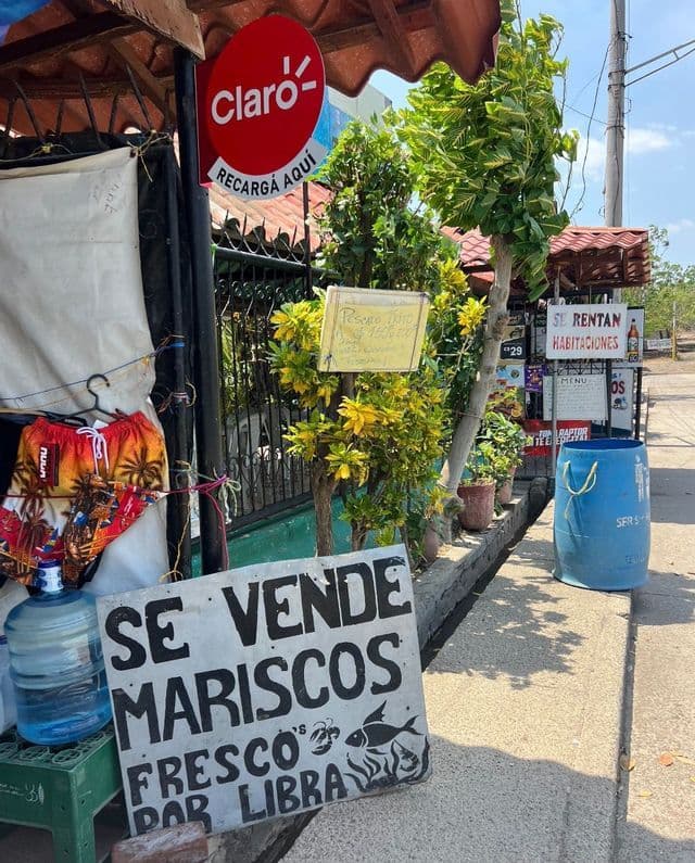 Ein handgemaltes Schild mit der Aufschrift 'Se Vende Mariscos' (Meeresfrüchte zu verkaufen) steht auf einem sonnigen Gehweg neben einem kleinen Laden am Straßenrand.