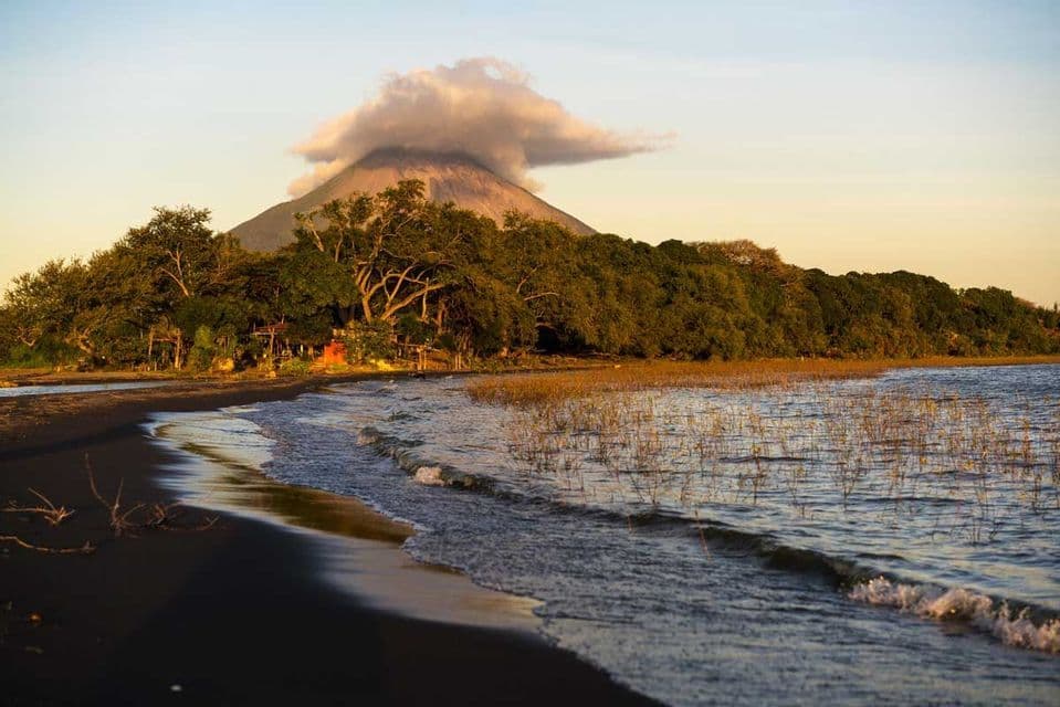 Ein Vulkan mit einer Wolke über seinem Gipfel erhebt sich hinter einem üppigen Wald, gesehen von einem dunklen Sandstrand mit sanft anrollenden Wellen.