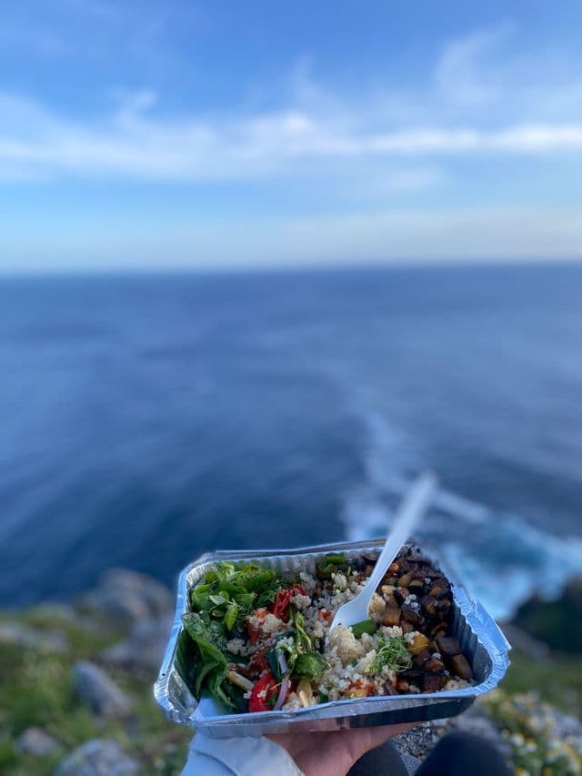 Una persona regge un contenitore di insalata da asporto con una forchetta, con vista sull'oceano da una scogliera sullo sfondo.