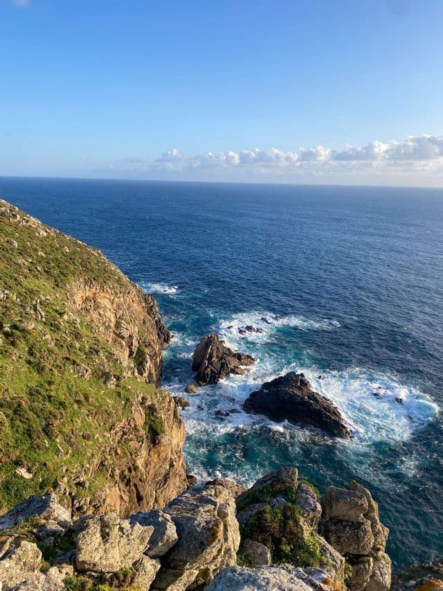 Una vista da un'alta scogliera erbosa a picco su un vasto oceano blu, con le onde che si infrangono sulle rocce sottostanti sotto un cielo parzialmente nuvoloso.